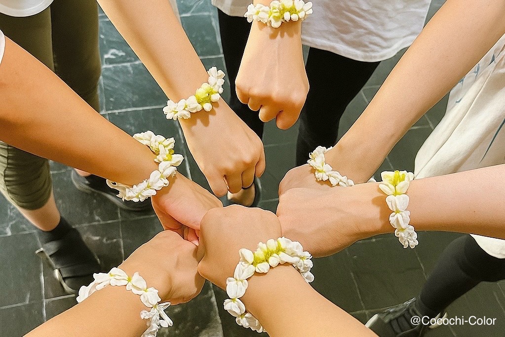 ルーシーダットンのクラス仲間とお揃いのジャスミンの花飾りをつけて手を合わせる様子。バンコクでの習い事の温かい雰囲気。
Ruesi Datton classmates wearing jasmine flower bracelets joining hands together — warm, friendly atmosphere in a Bangkok wellness class.